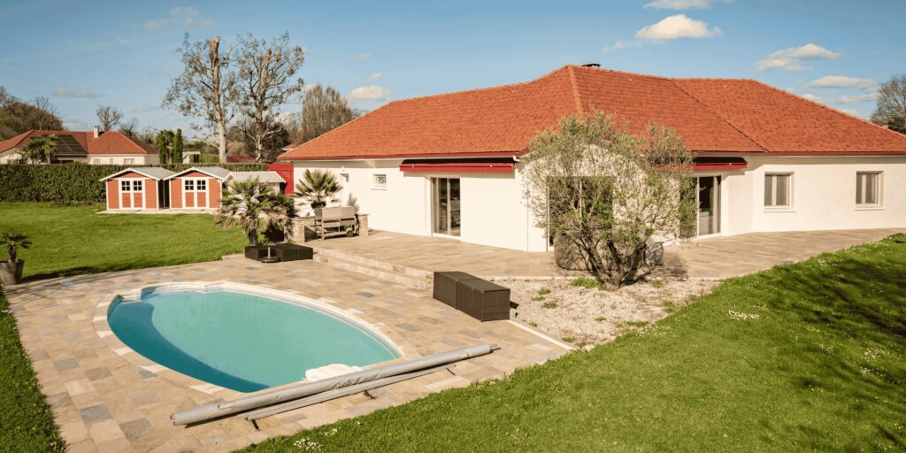 Terrasse et tour de piscine ovale en carrelage pierre beige devant une maison à Pau